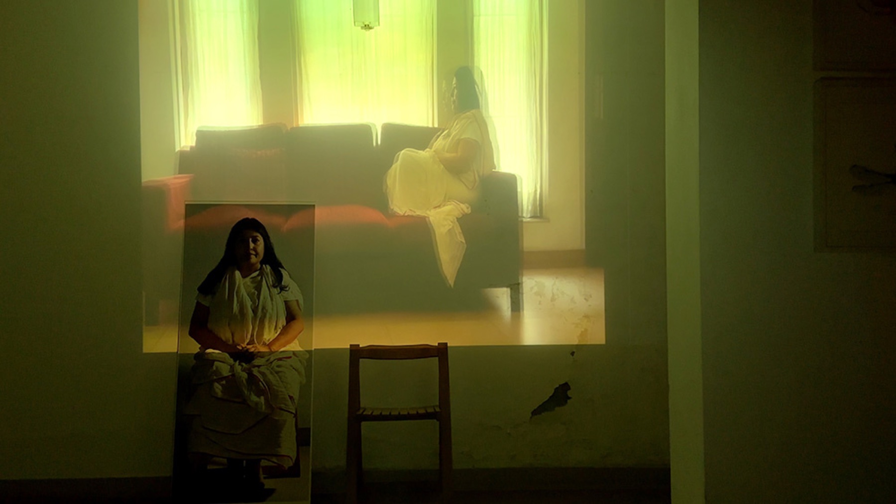 Projection of a seated figure and an empty wooden chair inside a domestic interior