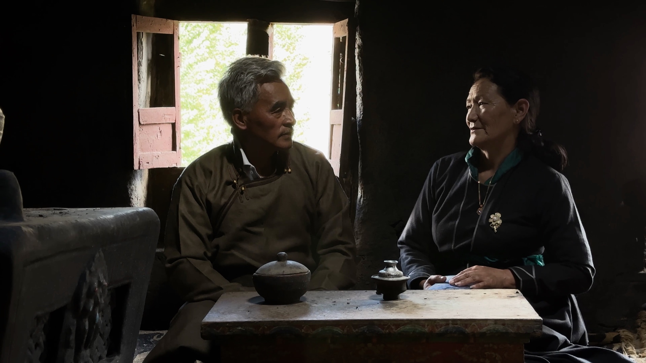 An elderly couple seated at a low table inside a lit interior in Ladakh